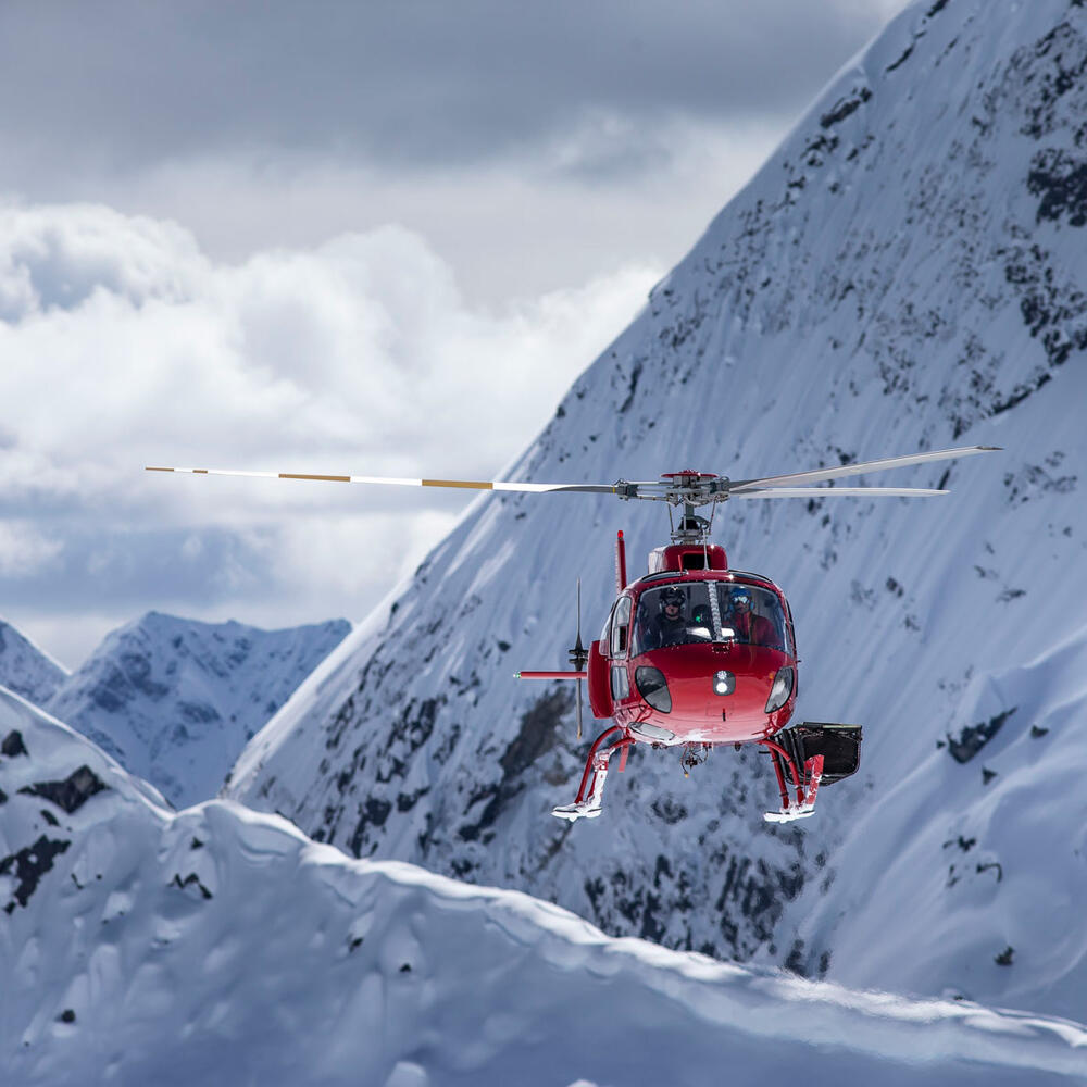 A Red Helicopter flying through a mountain valley covered in snow